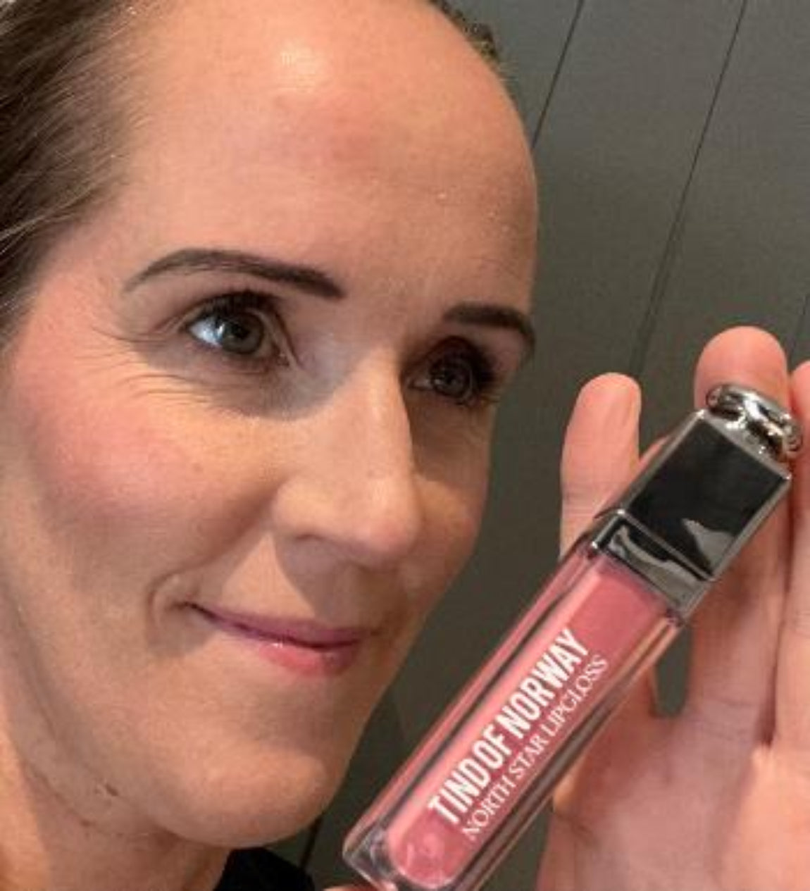 Kvinne med rosa farge på kinnene og blanke rosa lepper holder en rosa lipgloss fra Tind of Norway