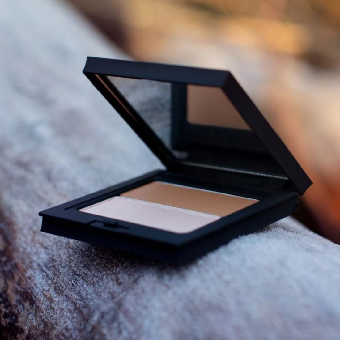 Åpen svart sminkepalett med speil, en lys highlighter og en brun sminkerefill på en frostdekket overflate