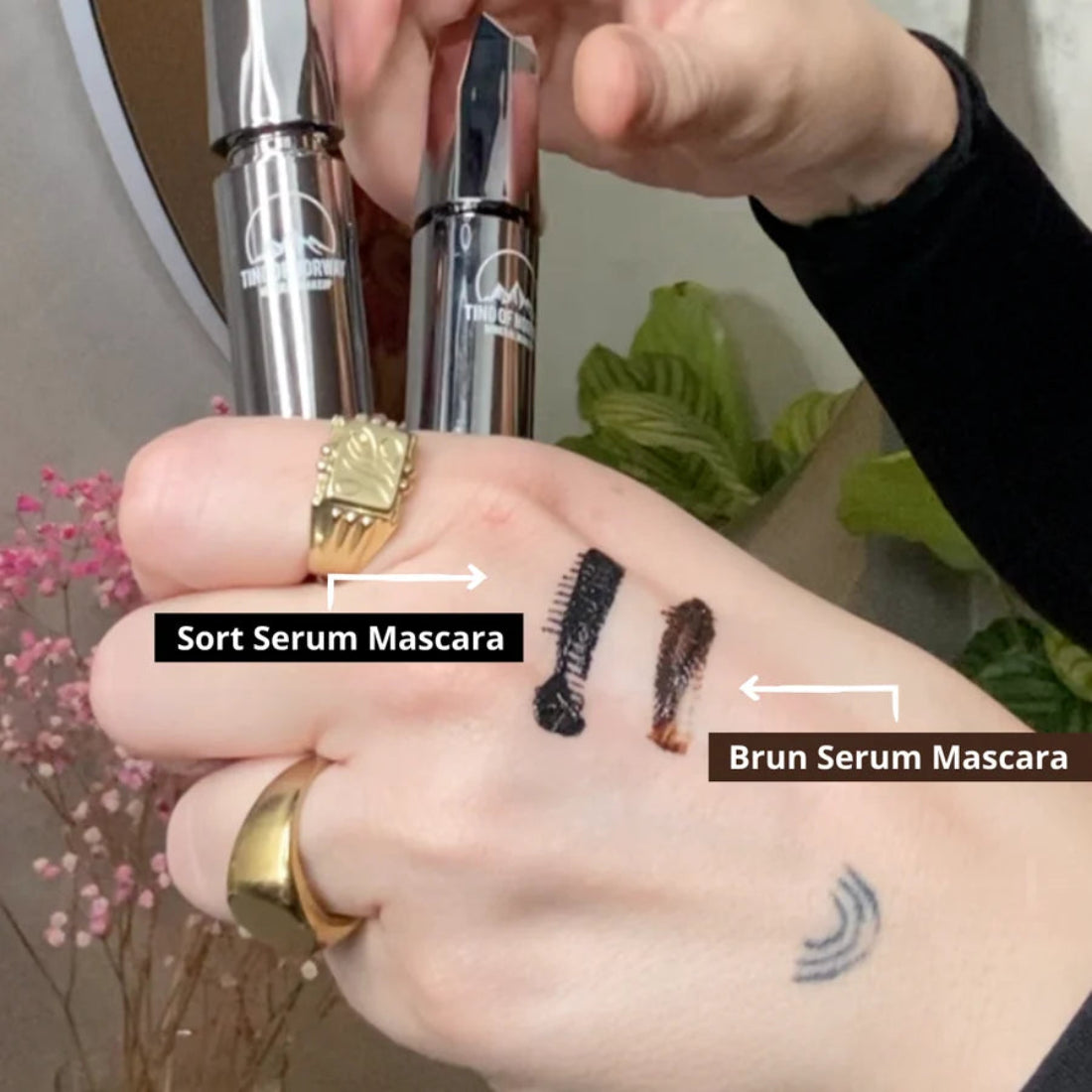 Tind of Norway Serum Mascara med sorte og brune fargeprøver på håndryggen og to sølvfargede mascarabeholdere
